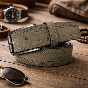 May include: Olive green suede belt with a dark metal buckle. The belt features a perforated design along the edges and a matching suede loop. Sunglasses and a watch are in the background. A stylish accessory.