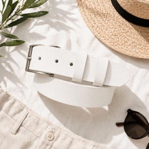 May include: A white leather belt with a silver buckle is displayed on a white surface. A straw hat with a black band and sunglasses are also visible. The scene suggests a summer fashion theme.