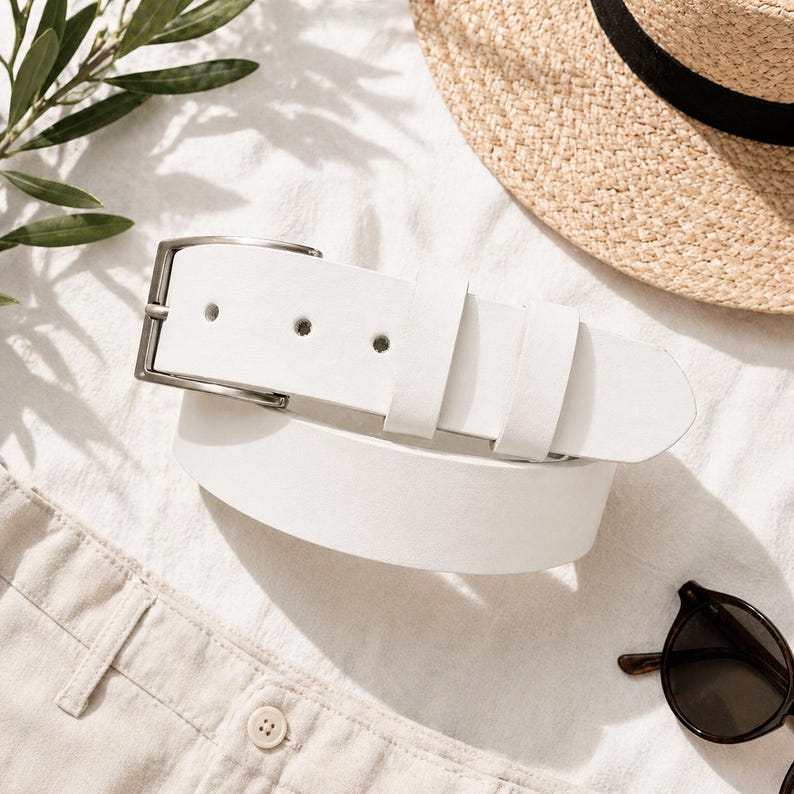 May include: A white leather belt with a silver buckle is displayed on a white surface. A straw hat with a black band and sunglasses are also visible. The scene suggests a summer fashion theme.