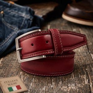 May include: A deep red leather belt featuring a pointed tip, white contrast stitching along the edges, and a brushed silver-tone metal buckle. A small square tag nearby displays the text ITALIAN MADE with a green, white, and red flag icon.