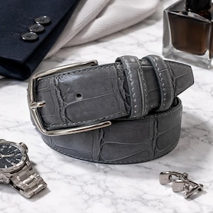 May include: A grey leather belt with a silver buckle, featuring a crocodile skin texture. The belt is displayed with a wristwatch, cufflinks, and a bottle of cologne, suggesting a theme of men's accessories.