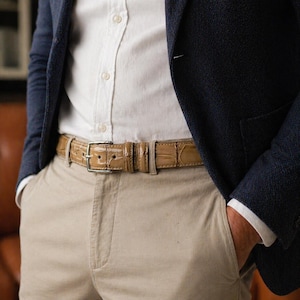 May include: A tan leather belt with a silver buckle is worn over beige trousers. The belt has a textured, crocodile-like pattern. A navy blue blazer and white shirt are visible in the background.