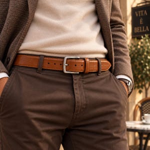 Può includere: Una cintura in pelle color cuoio con una sottile trama a griglia in rilievo e una fibbia in metallo argentato. La cintura è indossata sopra pantaloni marrone scuro, un maglione color crema e un blazer marrone a motivi. Sullo sfondo appare VITA E BELLA.