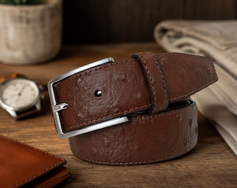 May include: A brown belt with a textured ostrich-like pattern and a silver-toned metal buckle. The belt features a single prong and a matching leather keeper loop, showcasing a distinct quill-follicle pattern across the surface of the strap.