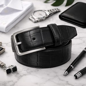 Puede incluir: Un cinturón de cuero negro con hebilla plateada y un patrón de cocodrilo texturizado. El cinturón está enrollado sobre una superficie de mármol, junto con un reloj, una cartera, una camisa, gemelos y bolígrafos.