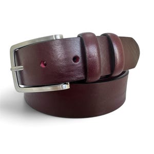 Può includere: Una cintura in pelle color bordeaux scuro con una finitura liscia e opaca e una fibbia in metallo color argento lucido. La cintura include due passanti in pelle abbinati e una serie di fori perforati per la regolazione, disposta a cerchio.