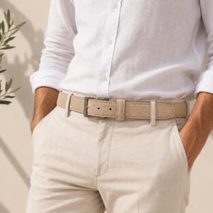 May include: A beige suede belt with visible stitching along the edges and a dark metal buckle, worn with a white long-sleeved button-down shirt and beige trousers. The belt features a classic design with a single prong and a matching leather loop.
