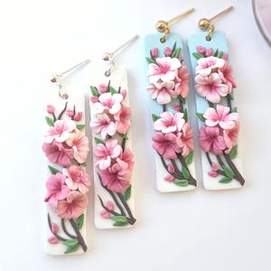 May include: Four pairs of floral dangle earrings. Each earring features a rectangular base with a 3D floral design of pink blossoms, green leaves, and brown stems. Two pairs have a light blue background, and two pairs have a white background.
