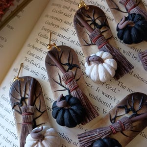 May include: A pair of handmade Halloween-themed earrings featuring a brown twig with a black and white pumpkin, a brown broom, and a black pumpkin. The earrings are attached to gold hooks.
