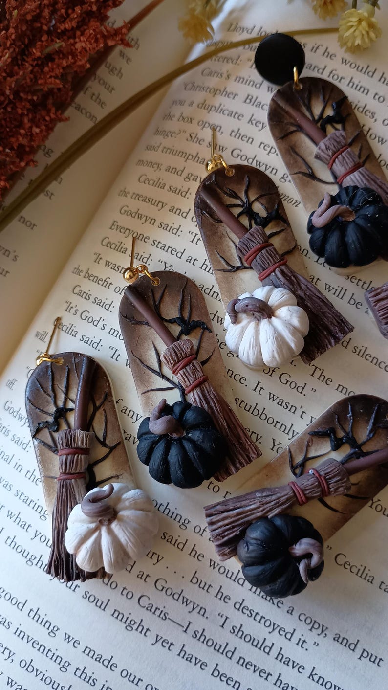 May include: A pair of handmade Halloween-themed earrings featuring a brown twig with a black and white pumpkin, a brown broom, and a black pumpkin. The earrings are attached to gold hooks.