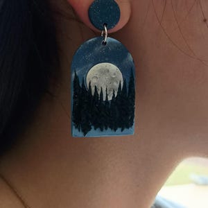 May include: A pair of dark blue polymer clay earrings with a full moon and forest silhouette design. The earrings are dangle style with a small round disc at the top.