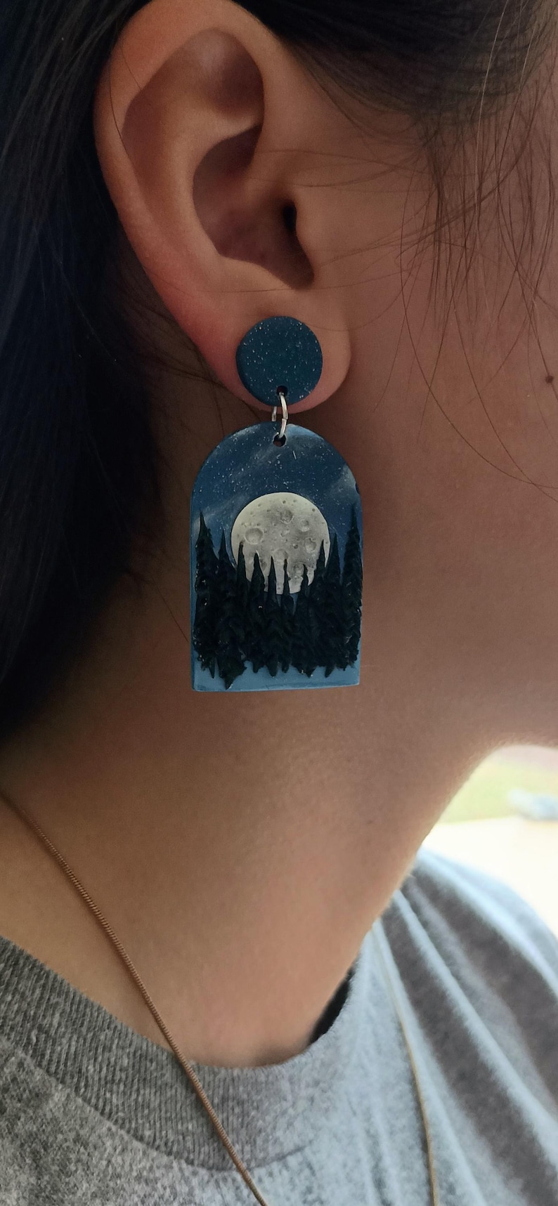 May include: A pair of dark blue polymer clay earrings with a full moon and forest silhouette design. The earrings are dangle style with a small round disc at the top.