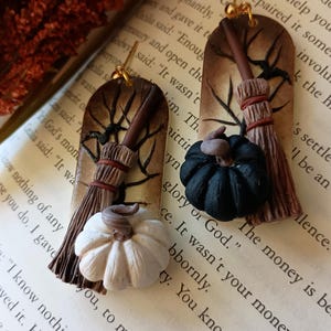 May include: A pair of handmade earrings featuring a brown and white broom with a black and white pumpkin. The earrings are on a book with text that reads "The money is be glory of God."