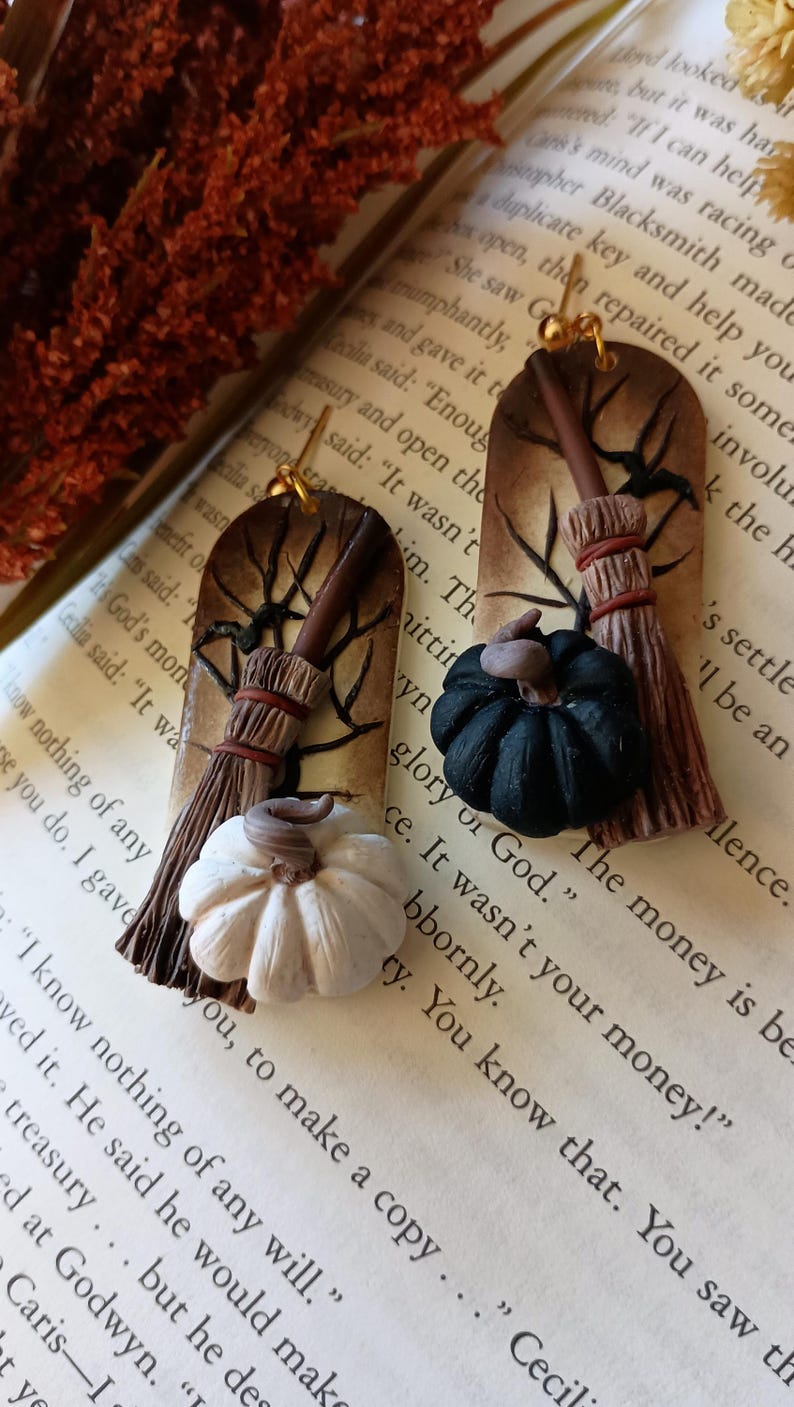 May include: A pair of handmade earrings featuring a brown and white broom with a black and white pumpkin. The earrings are on a book with text that reads "The money is be glory of God."