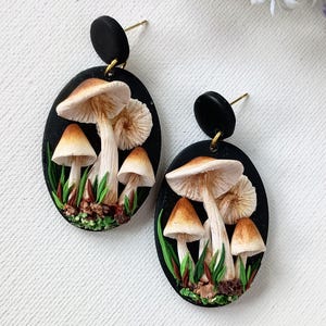 May include: A pair of black oval earrings with a mushroom design. Each earring features a cluster of white mushrooms with brown caps, set against a black background with green foliage.