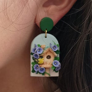 May include: A pair of polymer clay earrings featuring a blue and green birdhouse with a yellow bird and purple flowers. The earrings are hanging from green circles.