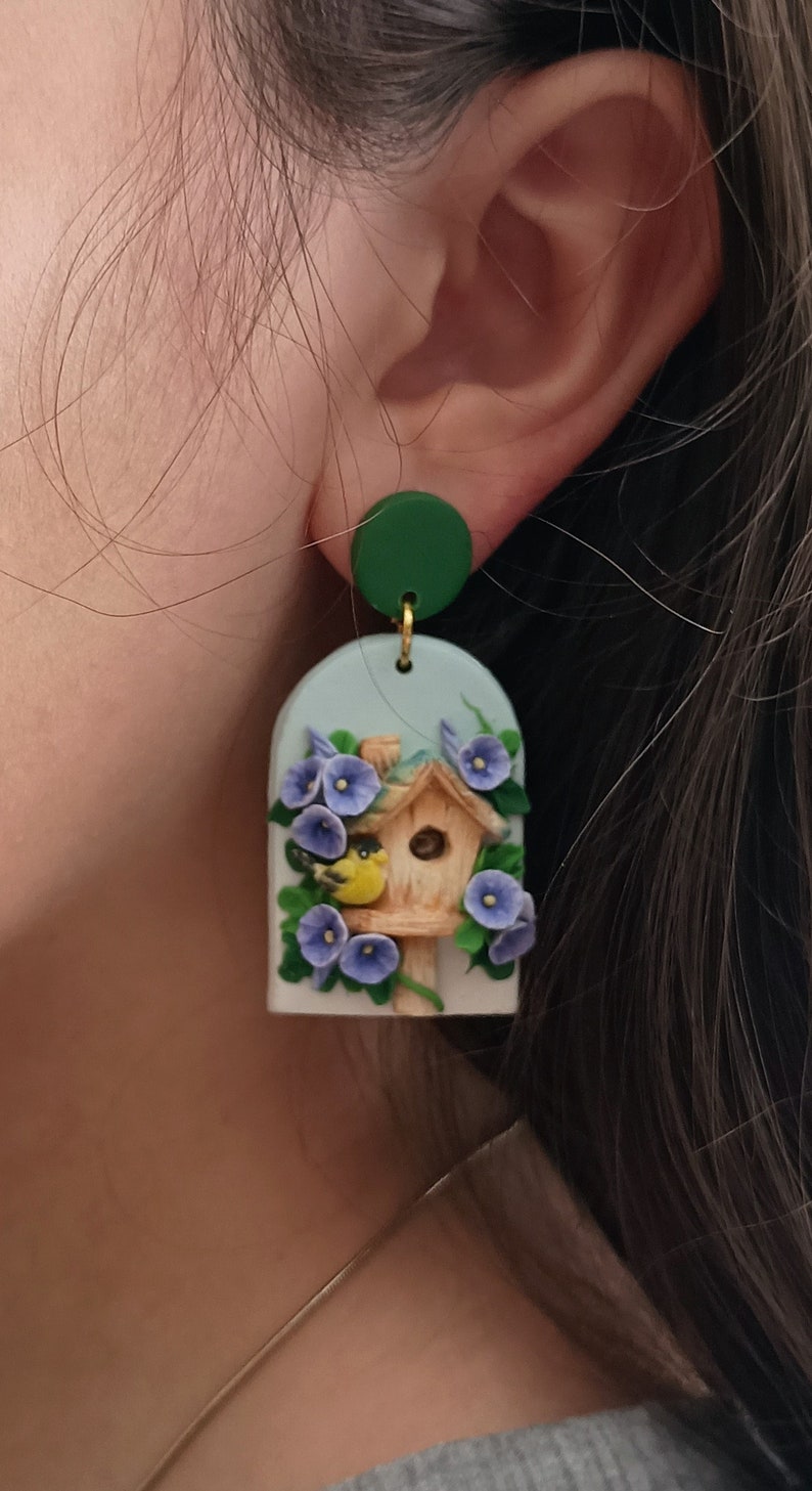 May include: A pair of polymer clay earrings featuring a blue and green birdhouse with a yellow bird and purple flowers. The earrings are hanging from green circles.