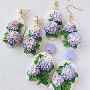 May include: Handmade polymer clay earrings with detailed hydrangea flower designs in purple, lavender, and white. The earrings have gold-coloured hooks and posts, and green leaves. The earrings are oval and arched shapes.