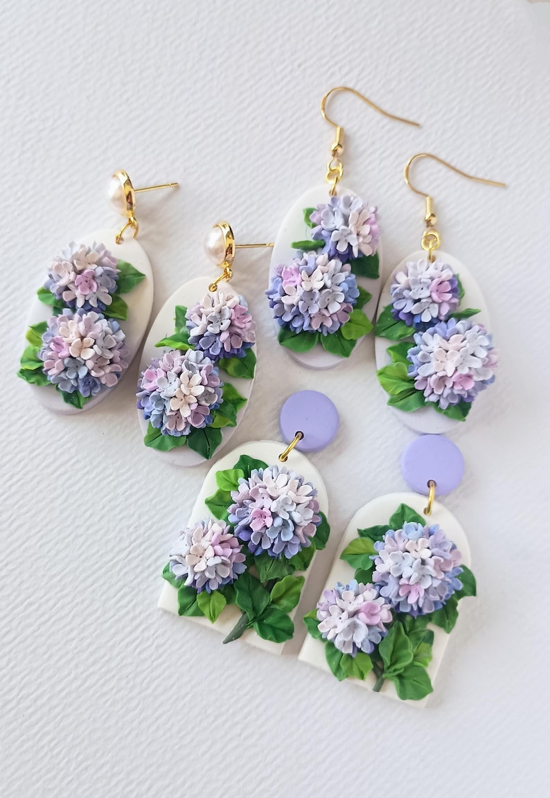 May include: Handmade polymer clay earrings with detailed hydrangea flower designs in purple, lavender, and white. The earrings have gold-coloured hooks and posts, and green leaves. The earrings are oval and arched shapes.