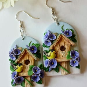 May include: A pair of handmade polymer clay earrings featuring a birdhouse with two yellow birds perched on the edge. The birdhouse is surrounded by purple flowers and green leaves. The earrings are attached to silver hooks.