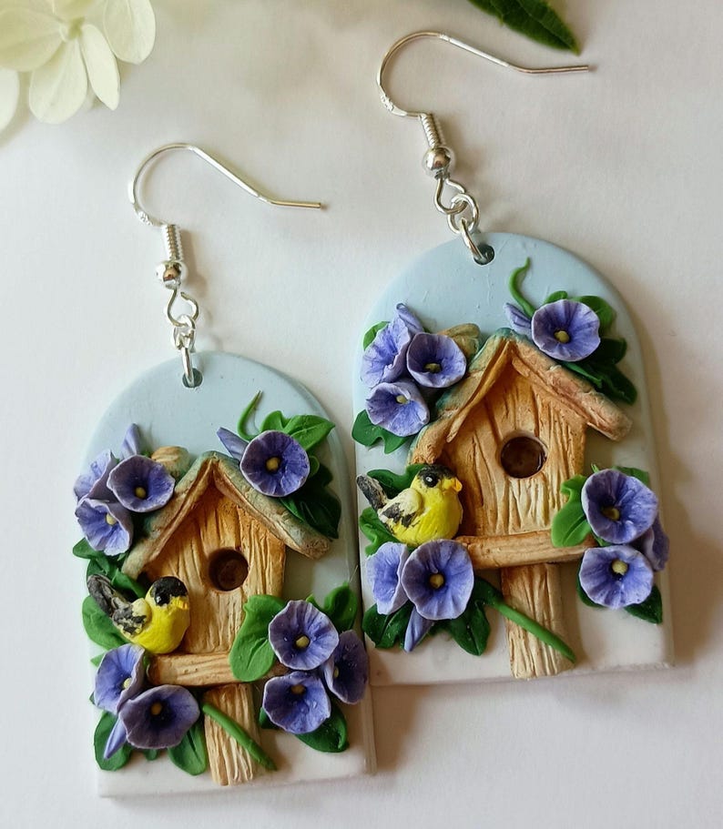 May include: A pair of handmade polymer clay earrings featuring a birdhouse with two yellow birds perched on the edge. The birdhouse is surrounded by purple flowers and green leaves. The earrings are attached to silver hooks.