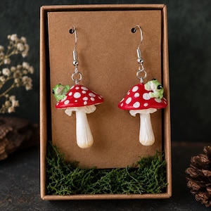 Peut inclure: Une paire de boucles d'oreilles pendantes représentant des champignons rouges à pois blancs, surmontés d'une petite figurine de grenouille verte. Elles sont suspendues à des crochets en métal argenté sur un support en carton brun.