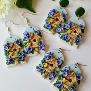May include: A pair of earrings featuring a birdhouse with two birds, surrounded by purple flowers. The birdhouse is made of brown clay and the flowers are made of purple clay. The earrings are attached to silver hooks.