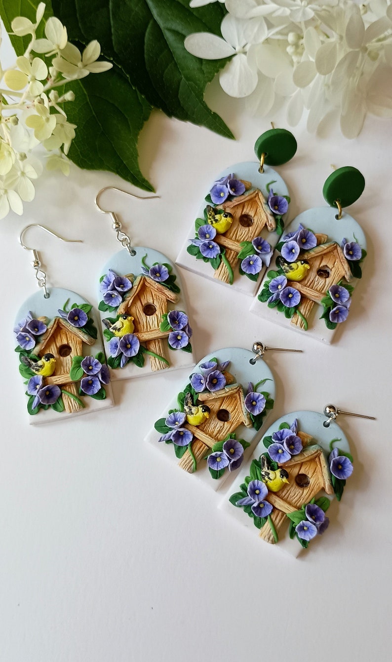 May include: A pair of earrings featuring a birdhouse with two birds, surrounded by purple flowers. The birdhouse is made of brown clay and the flowers are made of purple clay. The earrings are attached to silver hooks.