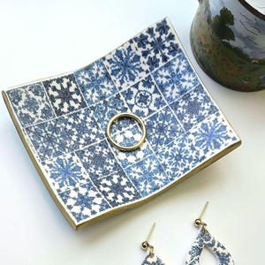 Op de afbeelding: Een vierkant keramisch sieradenschaaltje met een blauw-wit sneeuwvlokken tegelpatroon. Een gouden ring ligt in het midden. Goudkleurige randen omzomen het schaaltje. Een paar bijpassende oorbellen zijn rechtsonder zichtbaar.