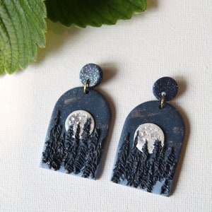 May include: A pair of blue and silver polymer clay earrings with a moon and forest scene. The earrings are shaped like an arch and have a glitter finish.