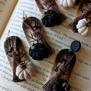 May include: A pair of Halloween-themed earrings featuring a brown and black broom, a black pumpkin, and a white pumpkin. The earrings are made of polymer clay and have gold hooks.