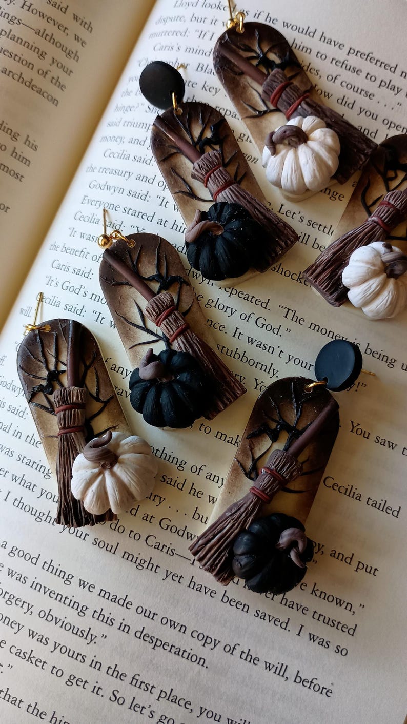 May include: A pair of Halloween-themed earrings featuring a brown and black broom, a black pumpkin, and a white pumpkin. The earrings are made of polymer clay and have gold hooks.