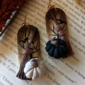 May include: A pair of handmade earrings featuring a black and white pumpkin and a broom with a brown handle. The earrings are made of polymer clay and are attached to gold ear wires.
