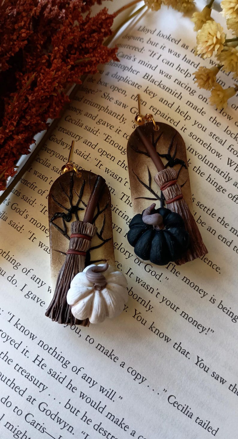 May include: A pair of handmade earrings featuring a black and white pumpkin and a broom with a brown handle. The earrings are made of polymer clay and are attached to gold ear wires.