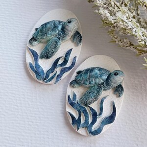 Puede incluir: Dos placas decorativas de forma ovalada con una tortuga marina tridimensional nadando sobre algas de color azul oscuro. Las tortugas est&aacute;n pintadas en tonos azules y verdes con caparazones texturizados sobre un fondo ovalado blanco roto.