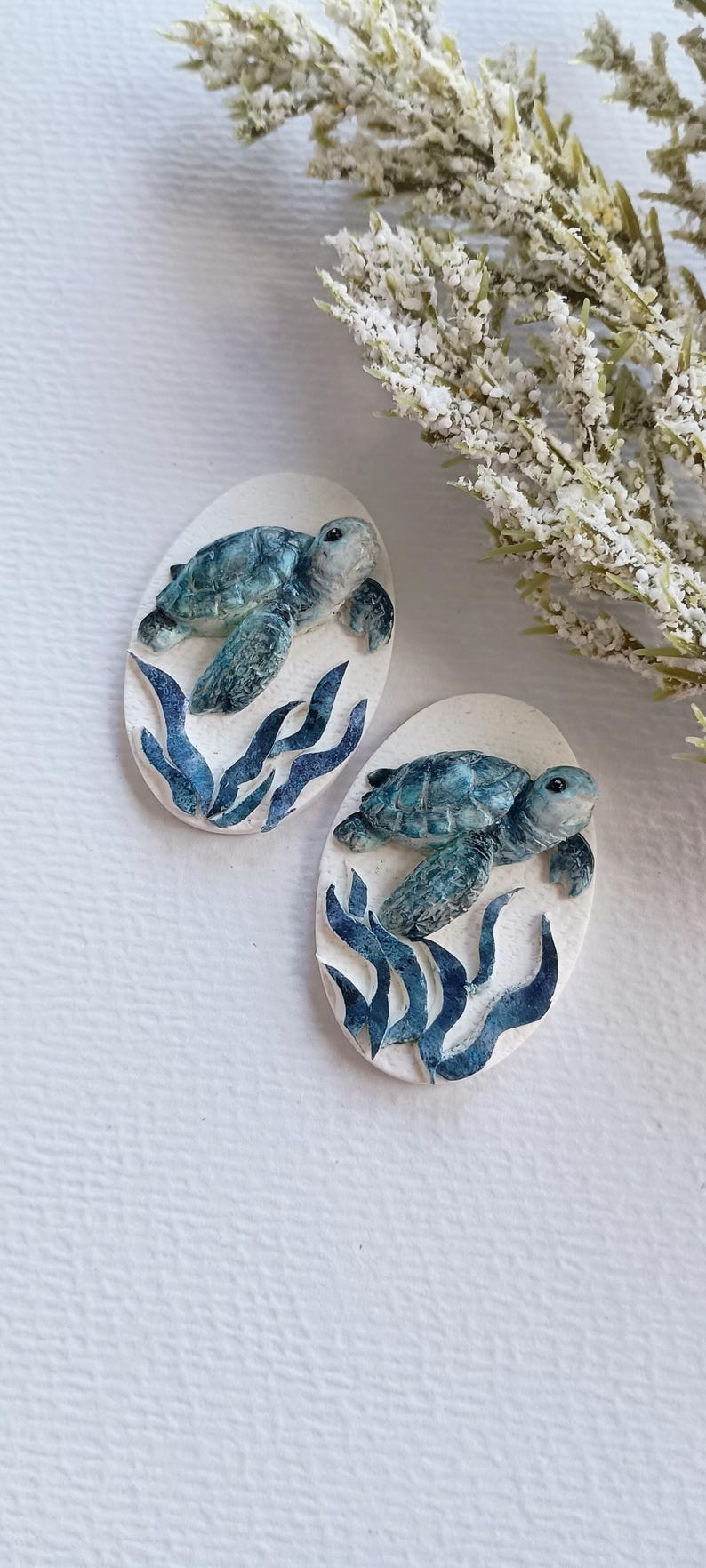 Puede incluir: Dos placas decorativas de forma ovalada con una tortuga marina tridimensional nadando sobre algas de color azul oscuro. Las tortugas est&aacute;n pintadas en tonos azules y verdes con caparazones texturizados sobre un fondo ovalado blanco roto.