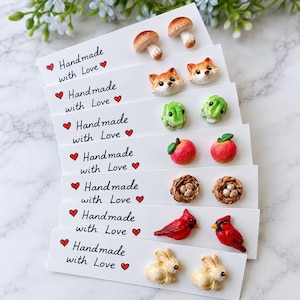 May include: Seven pairs of stud earrings on white cards labeled Handmade with Love. Designs include brown mushrooms, fox faces, green frogs, red apples, bird nests with eggs, red cardinal birds, and beige rabbits, all crafted in a miniature style.