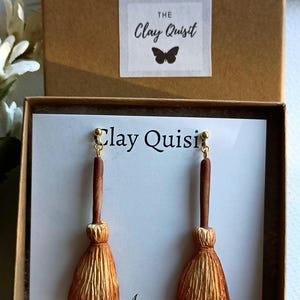 Puede incluir: Un par de pendientes de escoba marrones y dorados en una peque&ntilde;a caja con el texto "The Clay Quisit" en la tapa. Los pendientes se muestran en una tarjeta blanca con el texto "Clay Quisit" en ella.