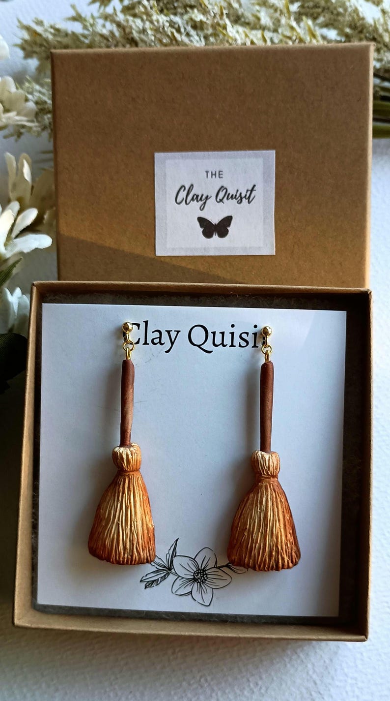 Puede incluir: Un par de pendientes de escoba marrones y dorados en una peque&ntilde;a caja con el texto "The Clay Quisit" en la tapa. Los pendientes se muestran en una tarjeta blanca con el texto "Clay Quisit" en ella.