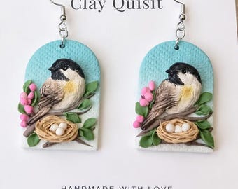 May include: A pair of dangle earrings featuring an arched design with a textured light blue background. Each earring displays a black-capped chickadee perched on a branch with pink berries and a small nest containing three white eggs. Clay Quisit.