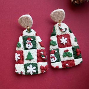 Op de afbeelding: Een paar feestelijke kerstoorbellen. Elke oorbel heeft een patchwork-ontwerp in wit, rood en groen met feestelijke elementen zoals sneeuwpoppen, sneeuwvlokken en zuurstokken. De oorbellen hebben een getextureerd, gebroken wit bovenstuk en goudkleurige haken.