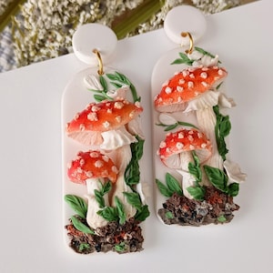 Op de afbeelding: Een paar witte keramische oorbellen met een paddenstoelontwerp. De paddenstoelen zijn rood met witte stippen en worden omringd door groene bladeren. De oorbellen zijn een grillig en door de natuur geïnspireerd accessoire.