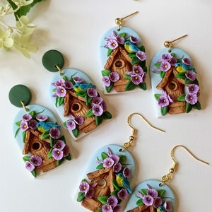 May include: A pair of handmade polymer clay earrings featuring a birdhouse with purple flowers and blue birds. The earrings are on a white background.