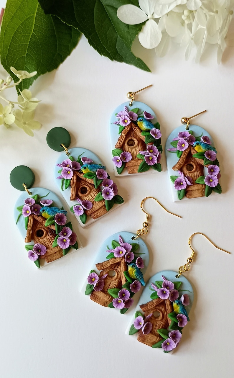 May include: A pair of handmade polymer clay earrings featuring a birdhouse with purple flowers and blue birds. The earrings are on a white background.