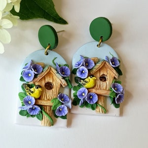 May include: A pair of handmade polymer clay earrings featuring a birdhouse with purple flowers and two yellow birds. The earrings are attached to green round discs.