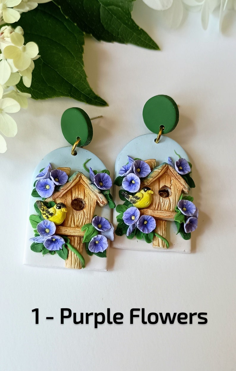 May include: A pair of handmade polymer clay earrings featuring a birdhouse with purple flowers and two yellow birds. The earrings are attached to green round discs.