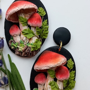 May include: A pair of black and red mushroom earrings. The earrings are made of polymer clay and feature a cluster of red mushrooms with white stems and green leaves on a black background.