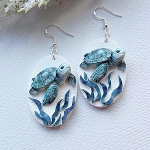 Puede incluir: Un par de pendientes colgantes ovalados con fondo blanco, un diseño en relieve de una tortuga marina azul y verde nadando sobre algas de color azul oscuro. Los pendientes están sujetos a ganchos de metal de color plateado.