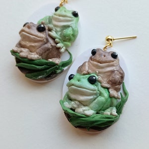 May include: A pair of frog earrings with a green frog on top of a brown frog. The frogs are sitting on a bed of green leaves. The earrings are made of polymer clay and have gold hooks.
