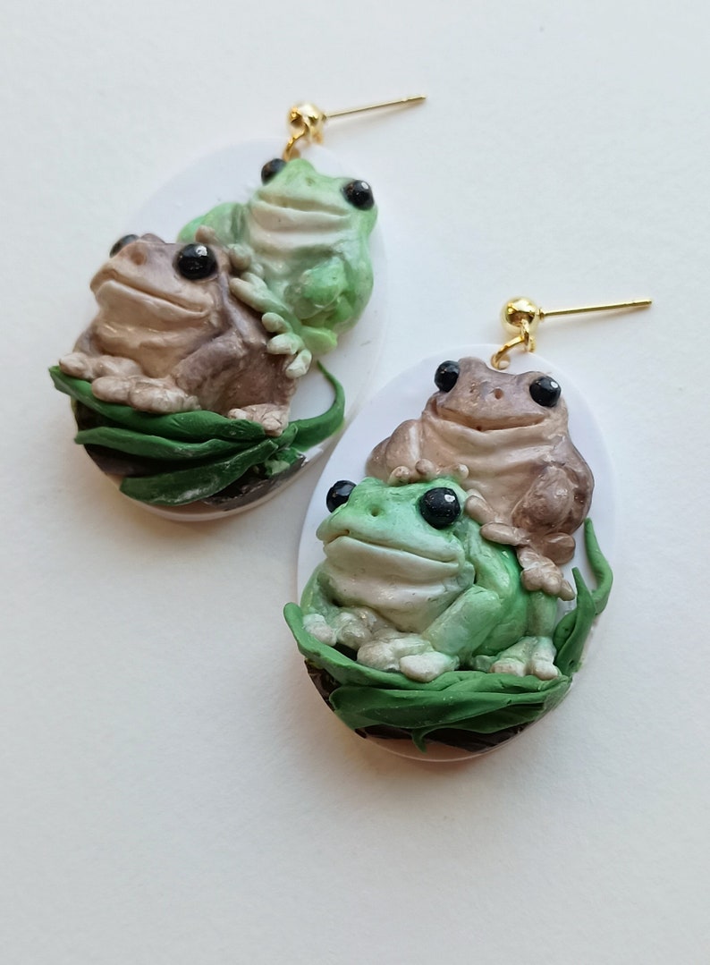 May include: A pair of frog earrings with a green frog on top of a brown frog. The frogs are sitting on a bed of green leaves. The earrings are made of polymer clay and have gold hooks.